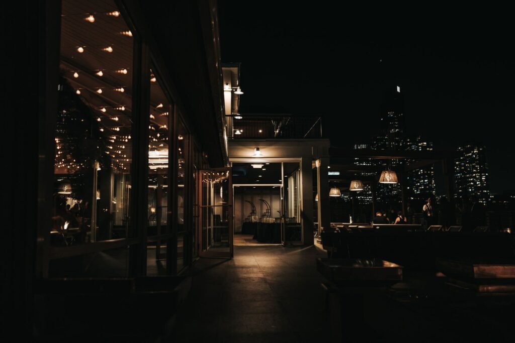 A rooftop bar terrace at night with warm lighting and a lit city skyline behind
