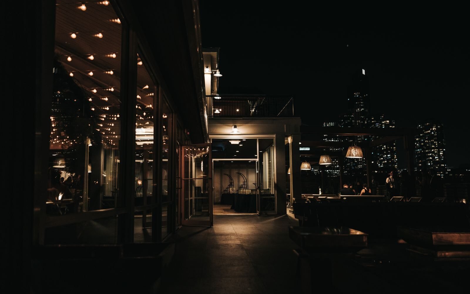 A rooftop bar terrace at night with warm lighting and a lit city skyline behind